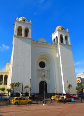 The Metropolitan Cathedral of San Salvador, an imposing white structure with twin towers, stands under a clear blue sky, partially obscured by a fence and bustling street activity.