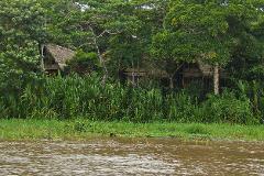 A river flows in front of lush green vegetation, with thatched-roof huts visible in the background.