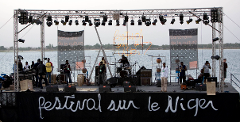 A band performs on a stage with lighting rigs and banners, on a body of water with greenery in the background. The stage reads "festival sur le Niger".