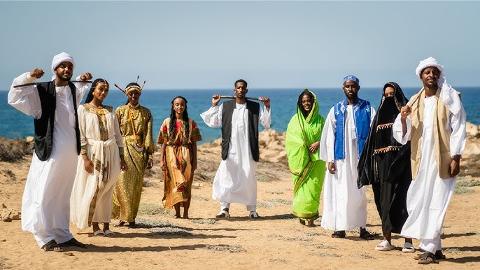A group of eight people in traditional Somali clothing are standing on a beach on a sunny day.