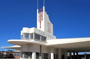 Certainly! Here's a description of the image:The Tagliero Service Station in Asmara, Eritrea, showcases striking Art Deco architecture with white curves, a distinctive tower featuring the Fiat logo, and long cantilevered wings, set against a clear blue sky.