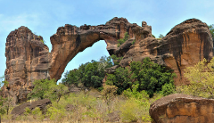 A natural sandstone arch dominates the landscape, connecting two large rock formations with lush green vegetation at its base.