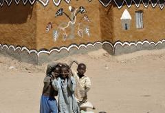 Three African children pose in front of a mud hut decorated with painted images of a tree, snake, and small house.