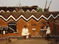 A colorful mural painted on an earthen wall depicts stylized scenes of village life in Burkina Faso.