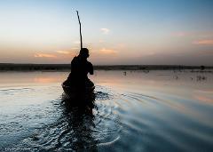 A silhouetted figure in a boat propels forward with a pole across a calm body of water, reflecting the colorful sunset sky.