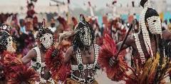 The image depicts people in traditional African tribal attire and masks, holding decorative fans, likely participating in a cultural dance or celebration.