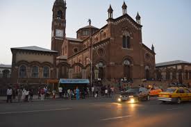 A red-brick cathedral with a tall clock tower stands behind a busy street scene filled with people and taxis.