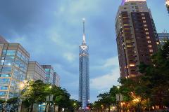 A tall, illuminated tower stands in the center of a cityscape with surrounding buildings, trees, and a cloudy sky at dusk.