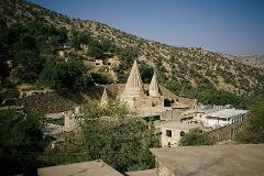 A serene view captures a stone building complex with three stepped pyramids in a mountainous, tree-dotted landscape under a clear blue sky.
