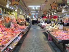 This is a bustling indoor market in Japan, filled with stalls displaying a variety of packaged foods, seafood, and snacks, under bright fluorescent lighting and colorful hanging signs.