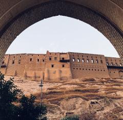 An old, tan brick castle sits atop a hill framed by a brick archway.
