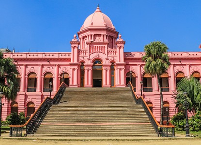 This is a photo of Ahsan Manzil, a pink palace with a grand staircase in Dhaka, Bangladesh, set against a clear blue sky and lush greenery.