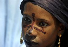A striking close-up of a woman with distinctive tribal face markings, a headdress, and golden earrings, likely taken during the Gerewol Festival.