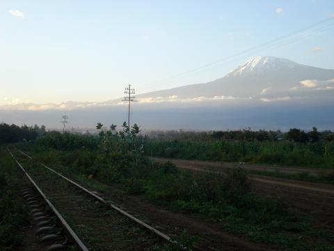 A scenic view of Mount Kilimanjaro behind a rural landscape with a railway track and vegetation.