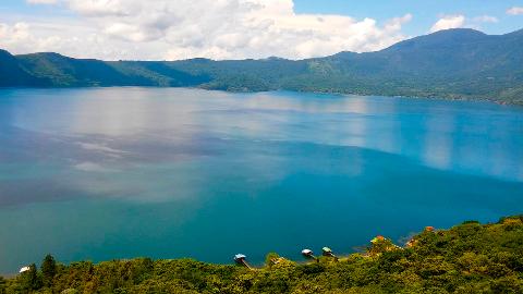 A scenic view of a large, blue lake surrounded by green mountains, with a lush, green shoreline in the foreground dotted with a few houses.