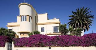 A striking cream-colored Art Deco-style house is partially hidden behind a vibrant wall of purple bougainvillea, with lush greenery and a palm tree adding to the tropical ambiance under a clear blue sky.