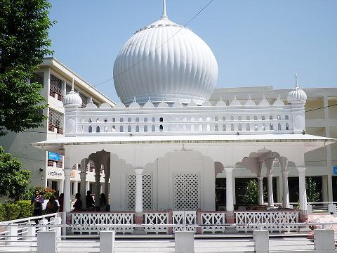 A bright, white, intricately designed temple with a large, rounded dome is front and center, surrounded by lush greenery and buildings under a clear, blue sky.
