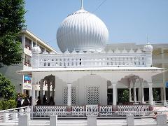 A bright, white, intricately designed temple with a large, rounded dome is front and center, surrounded by lush greenery and buildings under a clear, blue sky.