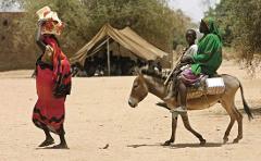 A woman with goods on her head walks past a donkey carrying two people in a rural setting with a tent and a group of people in the background.