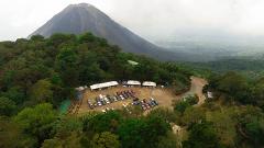 A high-angle shot showcases a parking lot nestled amidst lush greenery, with a towering, partly cloud-covered volcano dominating the backdrop.