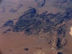 Certainly! Here's a description of the image:This aerial view shows the Aïr Mountains, a rugged and dark rock formation jutting out of the surrounding sandy desert landscape, showcasing their dramatic contrast in terrain. 