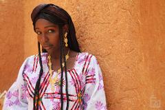 A striking portrait of a young woman with braids and a patterned top, adorned with gold jewelry, set against an earthen wall.