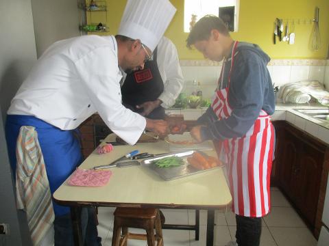 A chef and a young boy are preparing food together in a kitchen, with the chef wearing a white uniform and tall hat, and the boy wearing a red and white striped apron.