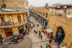An elevated view of a bustling street scene in Baghdad, Iraq, featuring people strolling along the pedestrian walkway with stalls selling books and other goods.