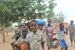 A group of happy African children are standing together, some smiling and waving, in a sandy outdoor setting with trees and simple buildings in the background.