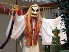 A figure in a white kimono and scary mask holds a ceremonial staff with outstretched arms in front of a decorated backdrop.
