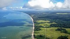 An aerial view shows a pristine coastline where the turquoise ocean meets a sandy beach, backed by lush green forests and rolling hills crisscrossed by dirt paths.