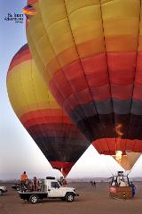 Two hot air balloons in the desert are being prepared for flight, with ground crew attending to them and a group of people in one of the balloon baskets.