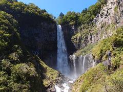 A majestic waterfall cascades down a rocky cliff surrounded by lush greenery under a clear blue sky.
