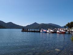 A picturesque view of a serene lake surrounded by lush mountains, featuring colorful swan-shaped paddle boats docked at a wooden pier under a clear blue sky.