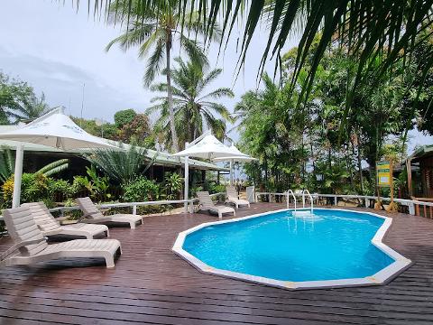 A tropical resort scene with a bright blue pool surrounded by a wooden deck, lounge chairs, palm trees, and lush greenery, under cloudy skies.