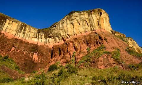 A striking rock formation with layers of reddish-brown and yellowish-tan rock under a clear blue sky, partially covered with vegetation.