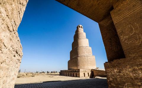 A spiral minaret towers against a clear blue sky, framed by the weathered brickwork of an ancient structure.