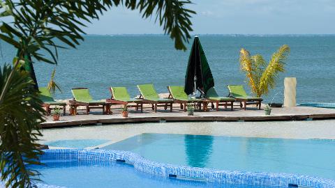 Loungers and umbrellas are set up on a wooden deck by a pool overlooking a calm blue sea.