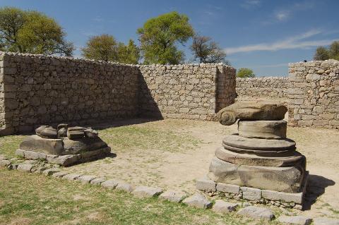 A courtyard with ruins of a stone wall and two crumbling, stone pillars, with some trees and a blue sky in the background.