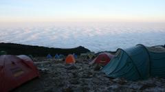 A campsite with several colorful tents sits above a blanket of clouds on Mt. Kilimanjaro, under a pale sky.