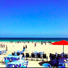 A sunny beach scene in Mogadishu features blue tables and chairs in the foreground, a crowded sandy beach with people enjoying the water, and a clear blue sky above.