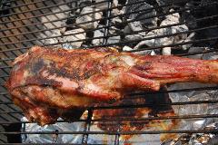 A grilled leg of lamb, well-browned, is placed on a wire grill over foil on a barbecue with visible coals above.