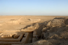 An aerial shot displays the ruins of an ancient structure situated in the middle of a vast, sandy desert with a clear blue sky on top.