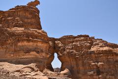 This eye-level shot showcases a massive, light brown sandstone rock formation with a natural archway under a clear blue sky.