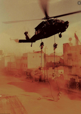 A Blackhawk helicopter hovers over a dusty urban landscape as soldiers descend on ropes toward the ground.