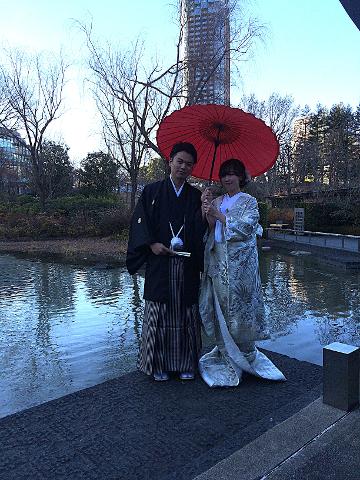 A traditionally-dressed Japanese couple poses by a pond under a red umbrella, with bare trees and a tall building in the background.