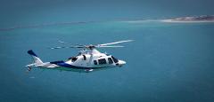 A white and blue helicopter flies over the ocean with an island in the background.