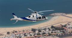 A white and blue helicopter flies over a beach with buildings and a circular structure in the background.