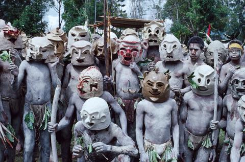 A group of men in Papua New Guinea, covered in light-colored paint and wearing elaborately decorated masks, are shown in a traditional gathering.