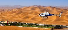 A blue and white helicopter flies over a sandy desert landscape with a road, trees, and a tent-like structure visible below.
